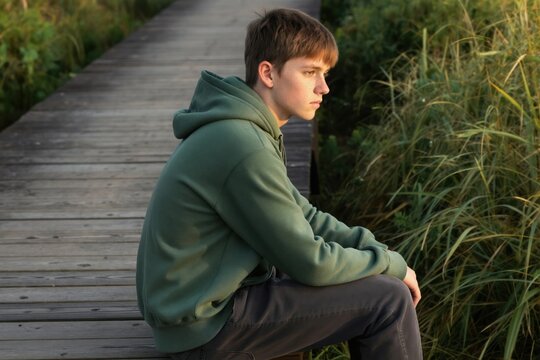 Young man sits on a wooden boardwalk in green hoodie. Boy looks thoughtfully at grassy nature. Teenager reflects in quiet solitude on path. Peaceful, outdoor mood.