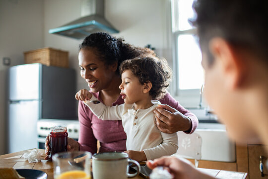 Mother helping toddler taste jam at home breakfast table