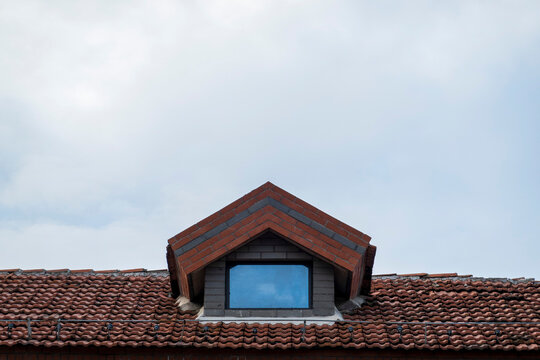 Minimalist architectural detail of a dormer window on a terracotta tiled roof under a pale sky. Sharp geometric lines and a cerulean reflection symbolize surveillance and residential isolation.