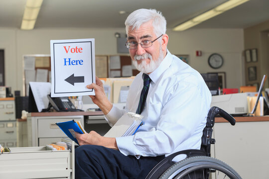Man with Muscular Dystrophy in a wheelchair working in his office and holding up a Vote Here sign
