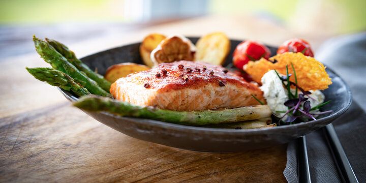 Grilled fish with fried bacon and green asparagus, served on black ceramic plate. Topped with potato wedges, garlic and szechuan pepper. Background for a healthy nutrion. Close-up. 