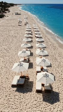 Beachside scene with rows of white umbrellas and lounge chairs on sandy shore, turquoise ocean waves gently lapping against the beach under clear blue sky