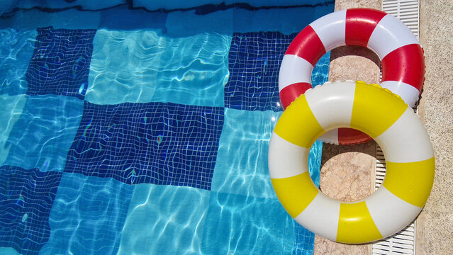 Summer vacation, travel, tourism concept. Red, white  and yellow striped  inflatable swim rings rest on the edge of a sparkling blue swimming pool.