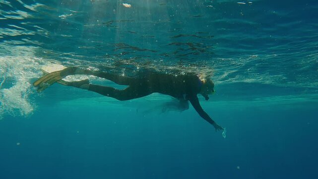 Female snorkeler, a cleanup volunteer picks up plastic debris drifting in turquoise water, covered with an oily layer on its surface, in bright sunlight, in backlit.