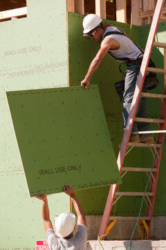 Hispanic carpenters pulling exterior sheathing up ladder at a house under construction