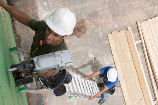 Hispanic carpenter using nail gun on top of ladder being steadied by another carpenter