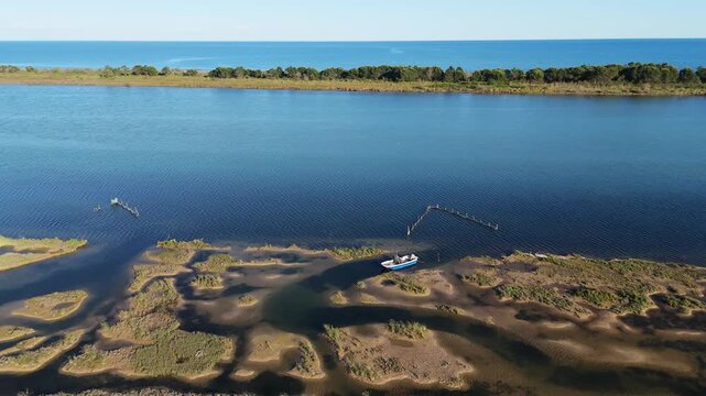 Drone aerial tracking shot over shallow tidal channels and textured mudflats, revealing a small lonely fishing vessel moored in the Venetian Lagoon near Eraclea at sunset, Italy