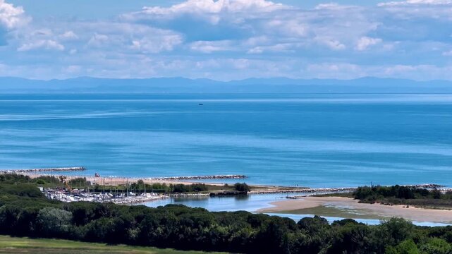 Wide aerial drone panorama of the Adriatic Sea coast near the Piave river mouth, revealing lush green shorelines and the distant Alps mountain range under blue sky, Eraclea, Italy