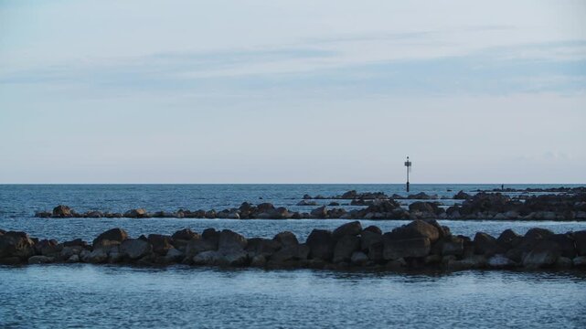 Calm maritime shot tracking along dark rocky breakwater barriers guarding the shallow shoreline entrance of the Venetian Lagoon during a soft twilight sunset near Eraclea, Italy