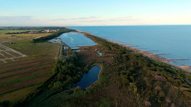 drone overflight following the narrow sand spit strip dividing the wild marshy wetlands of the Venetian Lagoon from the open sea waves during a soft golden sunset, Italy