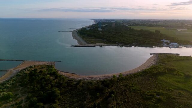 drone overflight of a curved sandy bay and coastal green forests, showcasing the dramatic meeting of land and calm sea waters at dusk near the Piave mouth, Eraclea, Italy