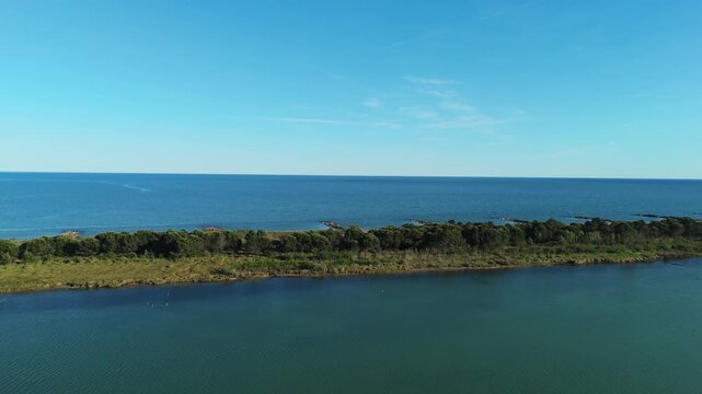 High wide drone panorama tracking the long sand spit strip separating the green lagoon marshlands from the calm sea waves at sunset near Eraclea and the Piave river mouth, Italy
