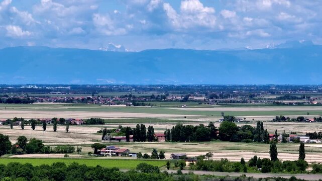 Wide aerial drone panorama of the fertile Venetian plain countryside with agricultural fields, looking towards the distant snowcapped Alps mountain range under blue sky, Italy