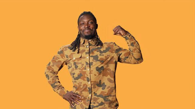 Athletic person flexing his arms muscles in studio and pointing up, showing off his strength and biceps standing over orange background, African American guy indicating upwards direction