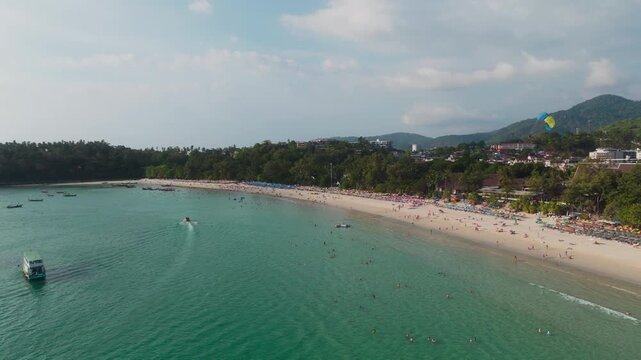 Drone follows parasail boat and tourist ferry over a turquoise Phuket bay.