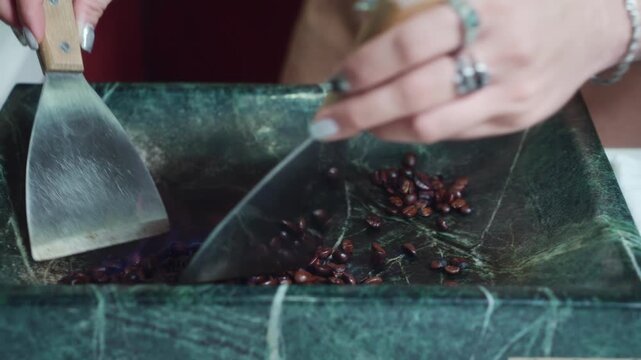 Close-up of barista hands using steel spatulas to toss coffee beans over a blue alcohol flambe flame inside a green marble plate. Premium artisanal roasting preparation