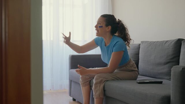 Young woman sitting on sofa reacting in anger to live sports game on TV at home. Female supporter in blue clothes and face paint passionate and involved screams and shouts in disappointment and stress