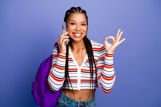Young chic student with braids on phone smiling and making ok gesture with purple backpack against blue background