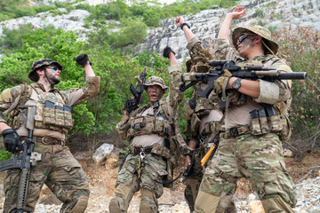 Fototapeta premium Group of four soldiers in camouflage uniform stacking hands during military tactical briefing outdoor, special forces operators showing team unity near rock cliff