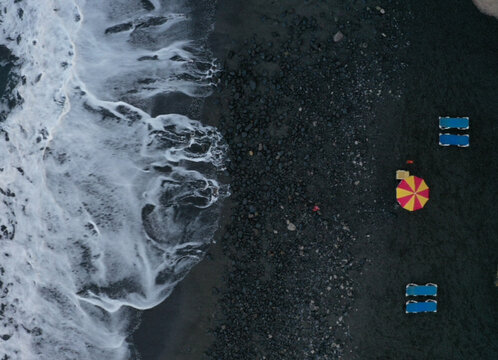 Aerial view of white waves crashing on a black sand beach with a colorful umbrella and blue sun loungers Acantilados de Los Gigantes, Canarias, Spain.