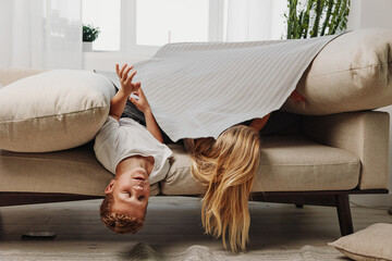 Obraz premium Two children play hiding under a blanket on beige sofa in bright living room, enjoying playful moment and family fun indoors together.