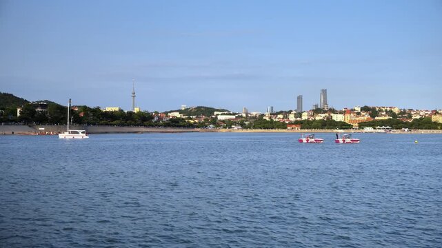 A calm coastal water scene with small boats, a distant urban skyline lined with trees, under a clear blue sky.