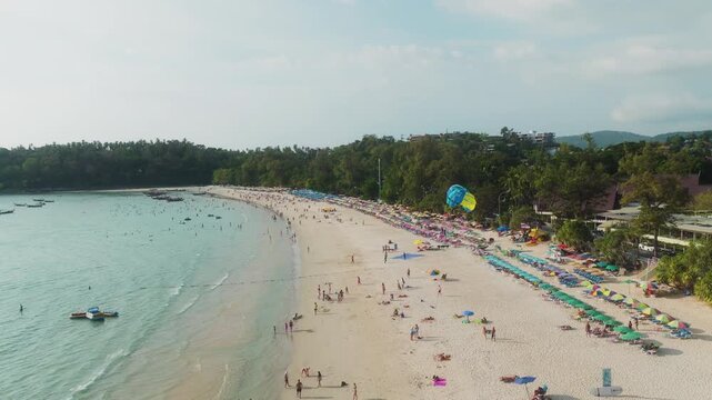 Low drone over a packed Phuket beach, parasail drifting above the crowd.
