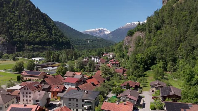 Aerial drone view of Rothenbrunnen, Switzerland, showing an alpine village surrounded by green hills, forested slopes, and mountain scenery in bright daylight.