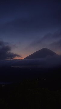 Vertical video. Dramatic Sunset Behind Mount Abang Above Lake Batur With Dark Storm Clouds and Distant Lights in Bali, Natural Beauty and Tranquility