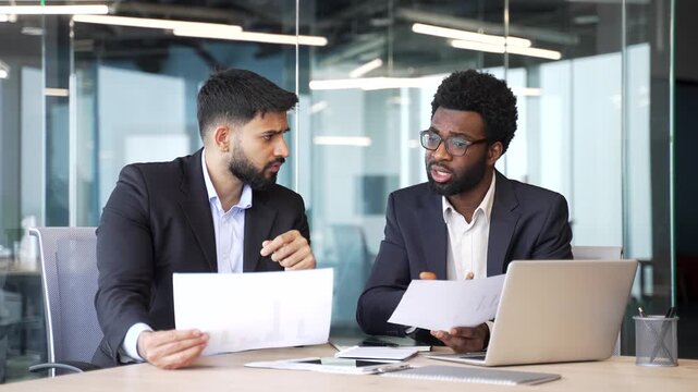 Two businessmen in formal suits sitting at office desk holding documents and discussing project. Multiethnic colleagues analyzing papers, planning strategy and exchanging ideas in corporate workspace.