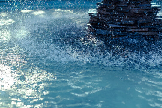 Close up of water falling from a stone tier fountain into a pool, creating droplets, splashes, ripples and reflections across the water surface. Abstract water feature background.