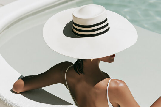 Woman in white wide brim hat with black stripes leaning at luxury pool edge in white swimsuit with dramatic shadow.