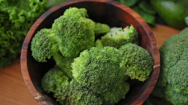 Fresh green broccoli florets in wooden bowl surrounded by leafy greens. Organic, high-quality natural products for healthy lifestyle, farm-raised vegetables. Slow motion