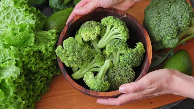 Woman's hands twirl wooden bowl of fresh broccoli florets surrounded by greenery. Concepts of healthy eating, organic vegetables, wholesome foods, healthy lifestyle. Slow motion