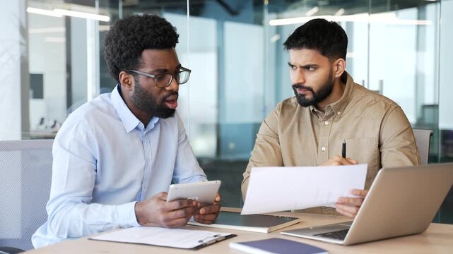 Two businessmen in casual clothes sitting at office desk reviewing document and talking. Multiethnic colleagues discussing project ideas, analyzing papers and collaborating on business tasks