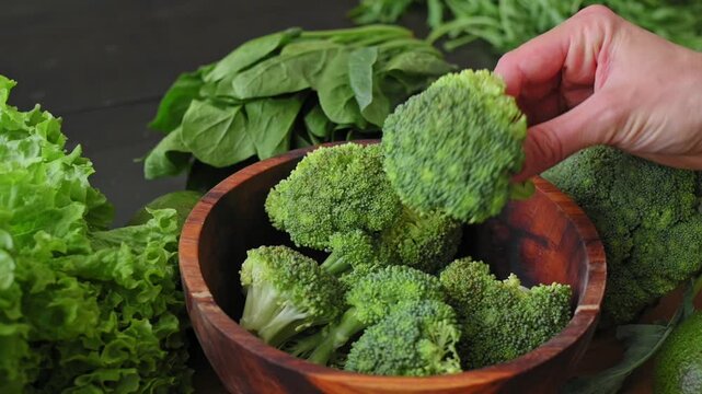Woman's hand picks up fresh broccoli floret from wooden bowl and twirls it, revealing the high-quality organic vegetable surrounded by lettuce and spinach leaves. Healthy food concept.