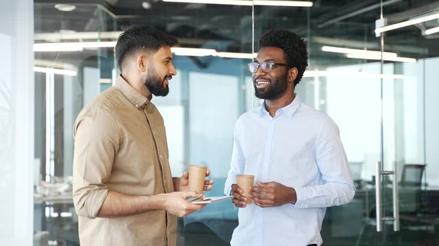 Two businessmen standing in office lobby with coffee cups and tablet, talking during break. Multiethnic colleagues in casual clothing sharing ideas, having friendly business conversation communicating