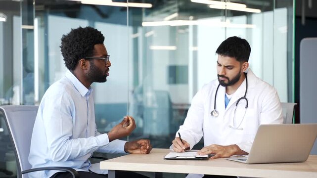 Doctor in white coat sitting at desk in hospital office talking with patient, listening carefully and writing notes. Male medical professional consulting and documenting information in modern clinic