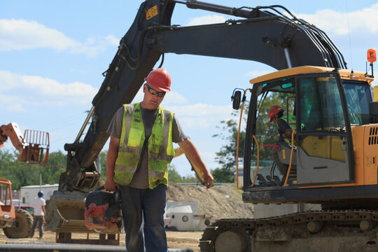 Construction engineer carrying pipe cutting saw at heavy construction site with excavator