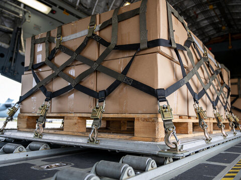 Cardboard boxes on a wooden pallet secured with nylon cargo netting inside a military transport plane.