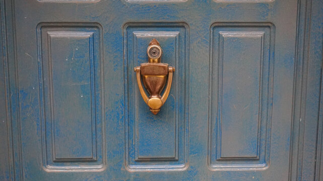 A close-up view of a blue painted wooden door featuring a classic brass door knocker with an integrated peephole centered on a vertical panel.