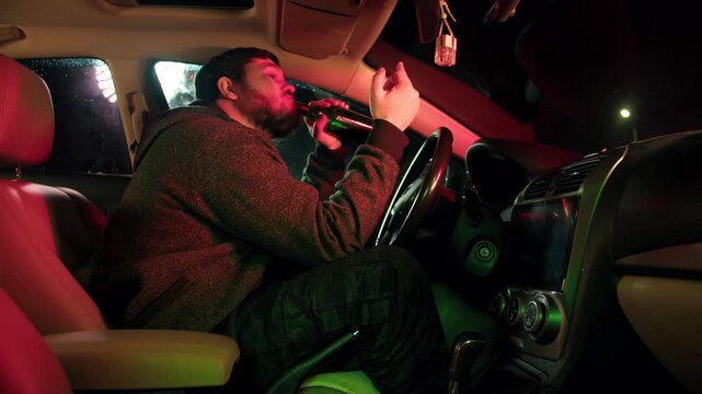 A drunken bearded caucasian man looks through the windshield of car, drinks alcohol from a bottle and ignores the police. Impudent driver shows the middle finger to law enforcement officials at night