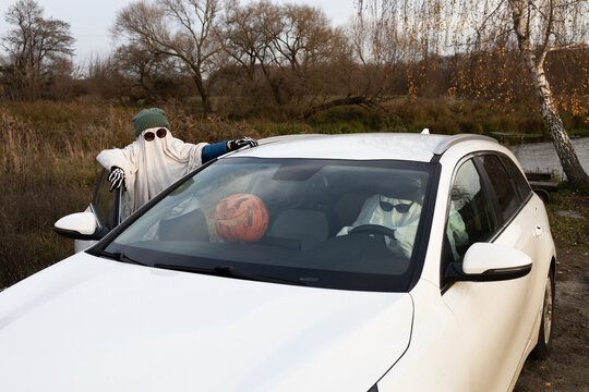 A child and an adult in white sheets and black sunglasses are driving to a party in a white car. People in ghost costumes. A funny Halloween photo. Positive moments of the holiday