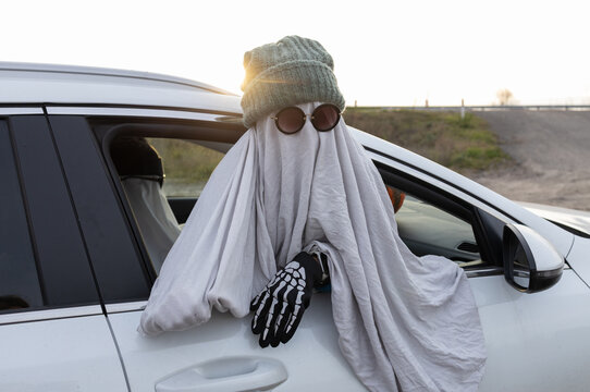 An unrecognizable child in a white blanket, hat, and black glasses looks out the window of a white car. People in Halloween ghost costumes are driving to a party. View from the window.