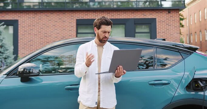 Adult man standing near a car on a city street, holding a tablet and looking at the screen with frustration