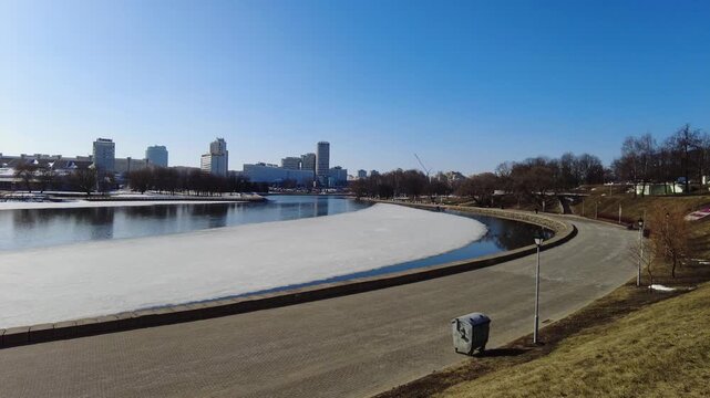 People walk along the riverbank on a sunny day. The river shows patches of ice as city buildings stand in the background. It is early spring and weather is clear.