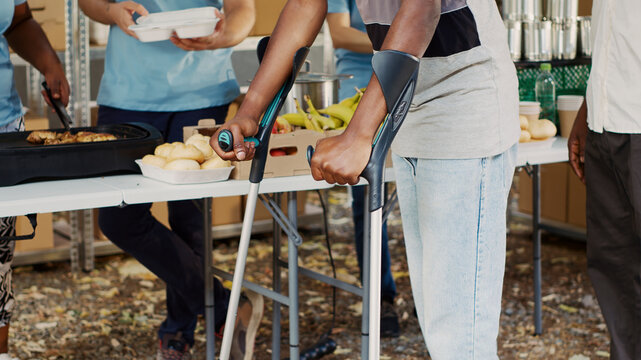 African American woman volunteer assists and supports crippled man at outdoor food bank, with the hungry, poor and homeless people receive hunger relief assistance from multiethnic voluntary.