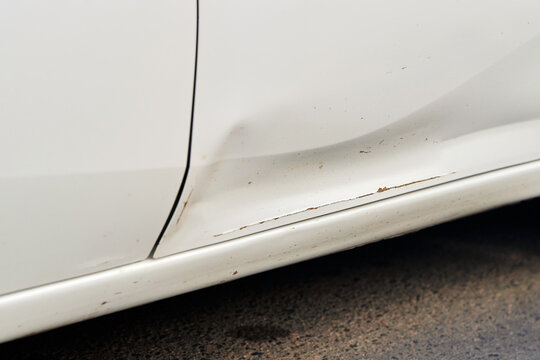 Scraped white car body with visible rust formation, paint damage on lower door section. Vehicle door panel with mechanical collision impact damage, sharp dent and exposed rusty metal. Selective focus