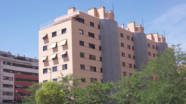 Modern residential building with beige facade and gray accents. Several windows and retractable awnings visible. Trees with green foliage frame the lower portion. Clear blue sky with no clouds