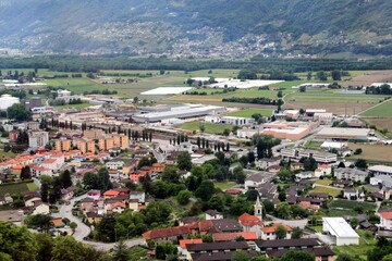 Fototapeta premium Aerial view of a vibrant town with red rooftops, church tower, railway yard, and green mountains blending urban life with natural landscapes.